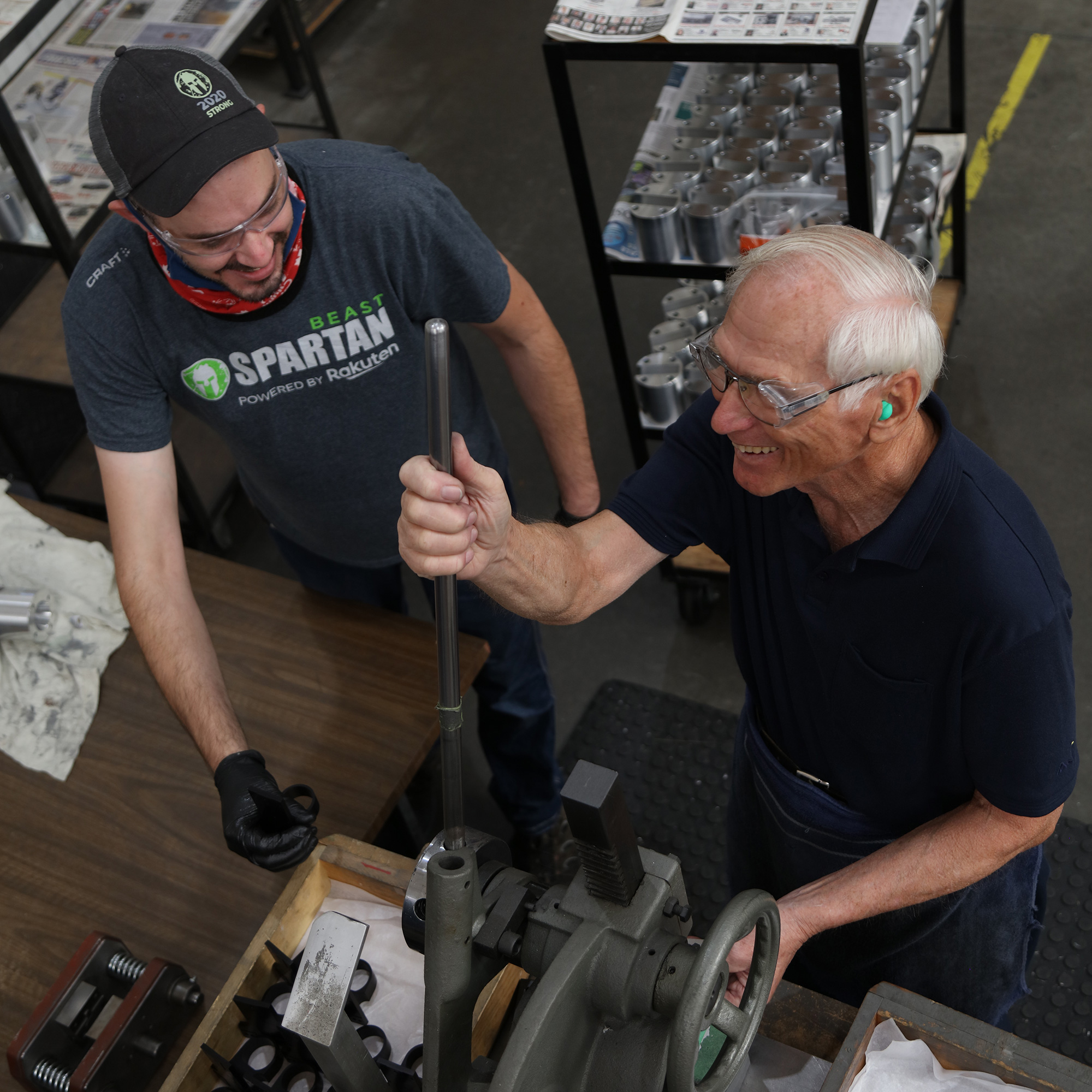 an employee works at a machine, smiling, while a coworker observes him