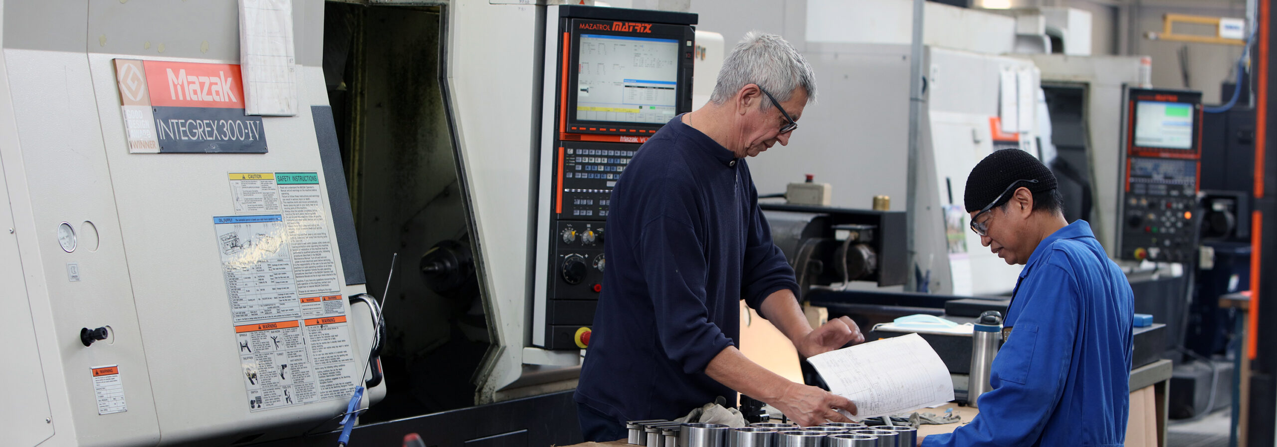 two employees stand at a table with components on it, looking at a piece of paper